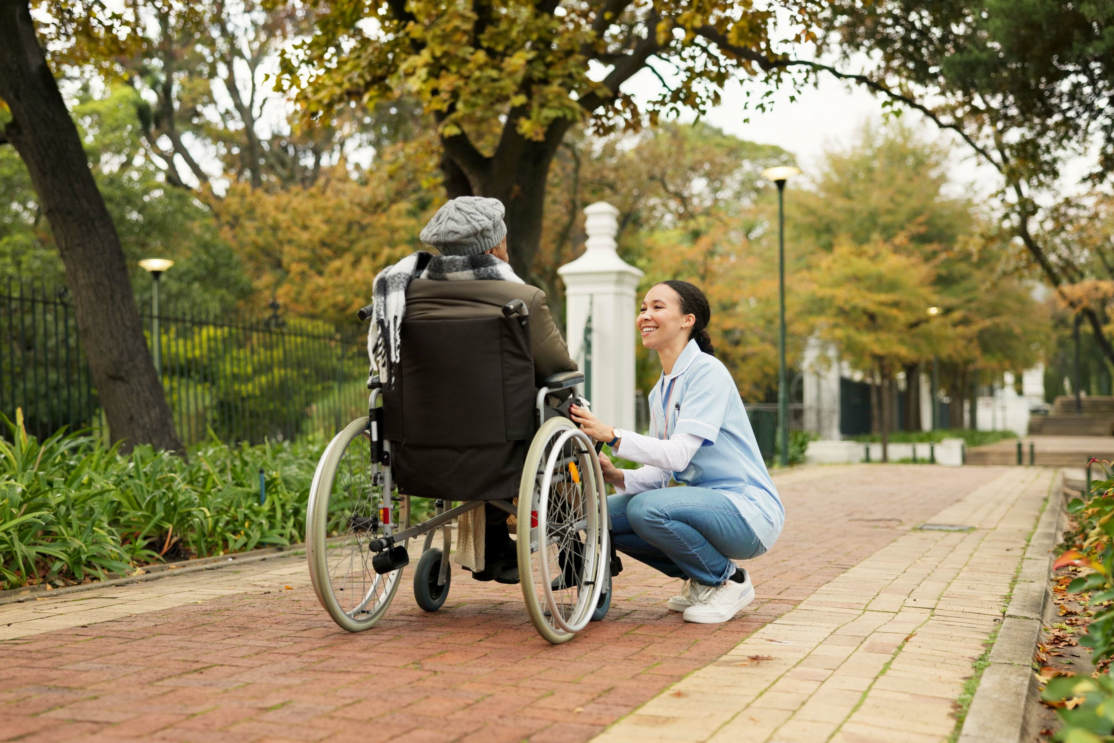 A healthcare worker assisting an elderly wheelchair user on an accessible, tree-lined park pathway.