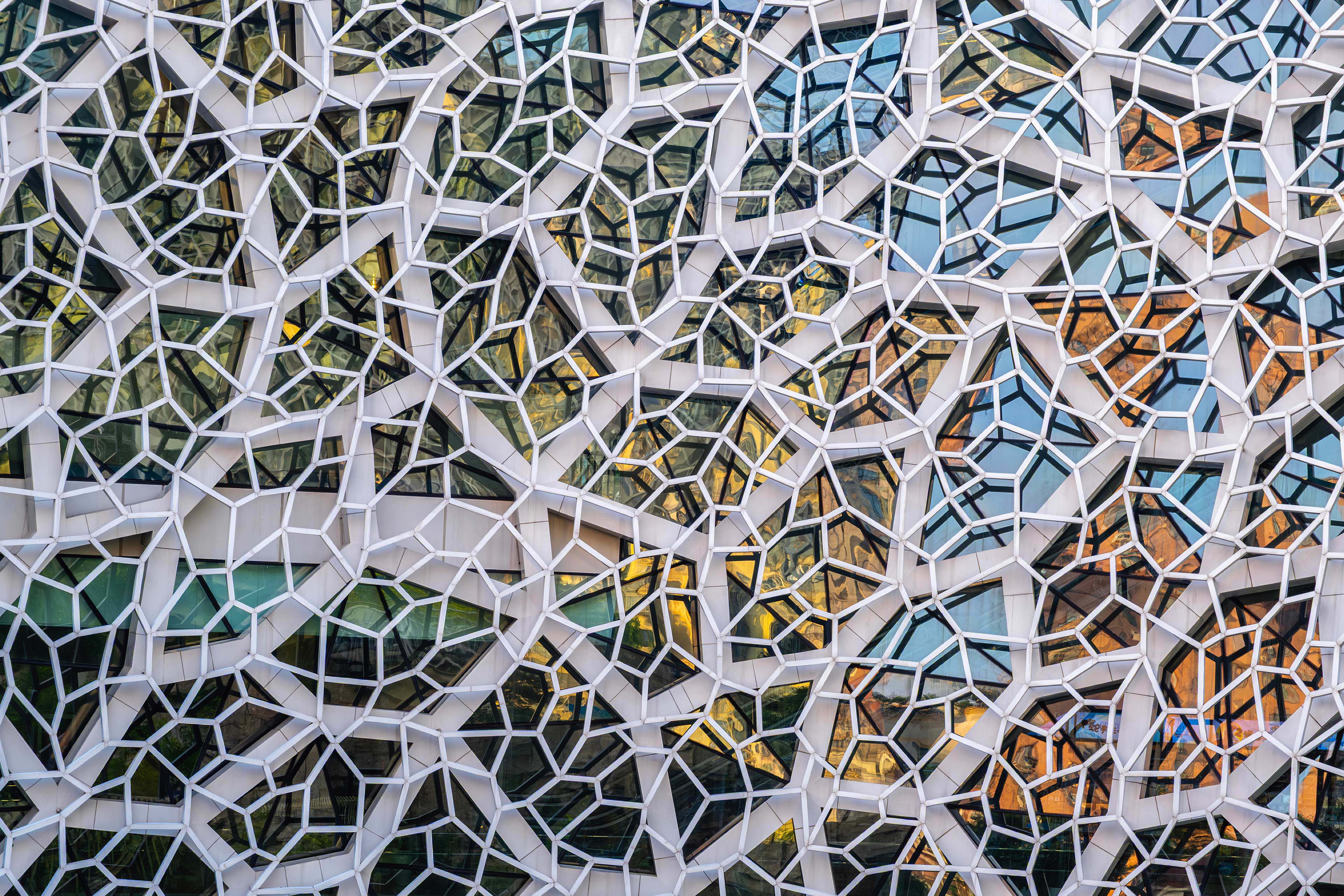 Modern facade with white hexagonal and irregular frames enclosing glass panels in a geometric mosaic pattern.