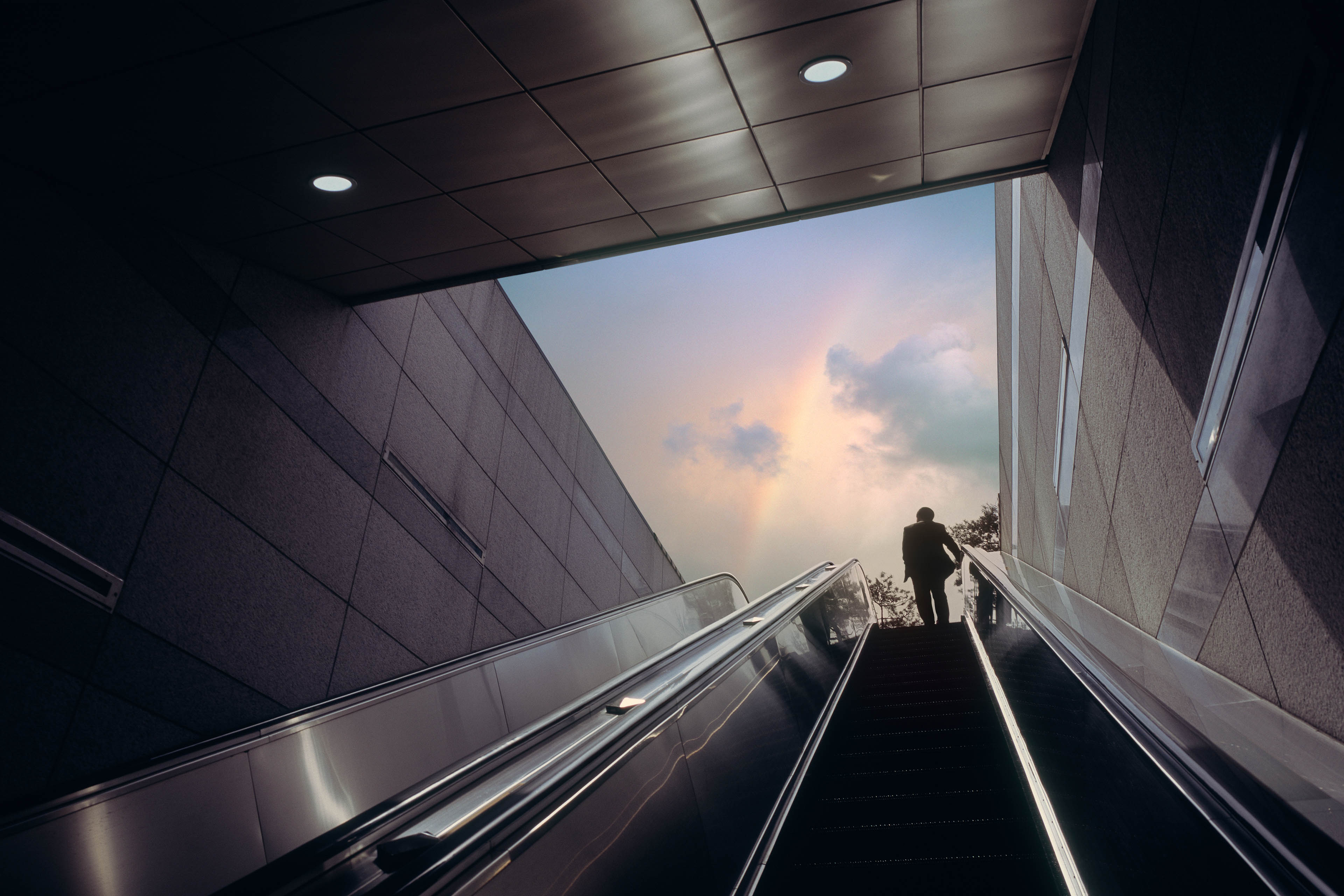 Silhouette sur un escalator montant vers un ciel éclairé par un arc-en-ciel.