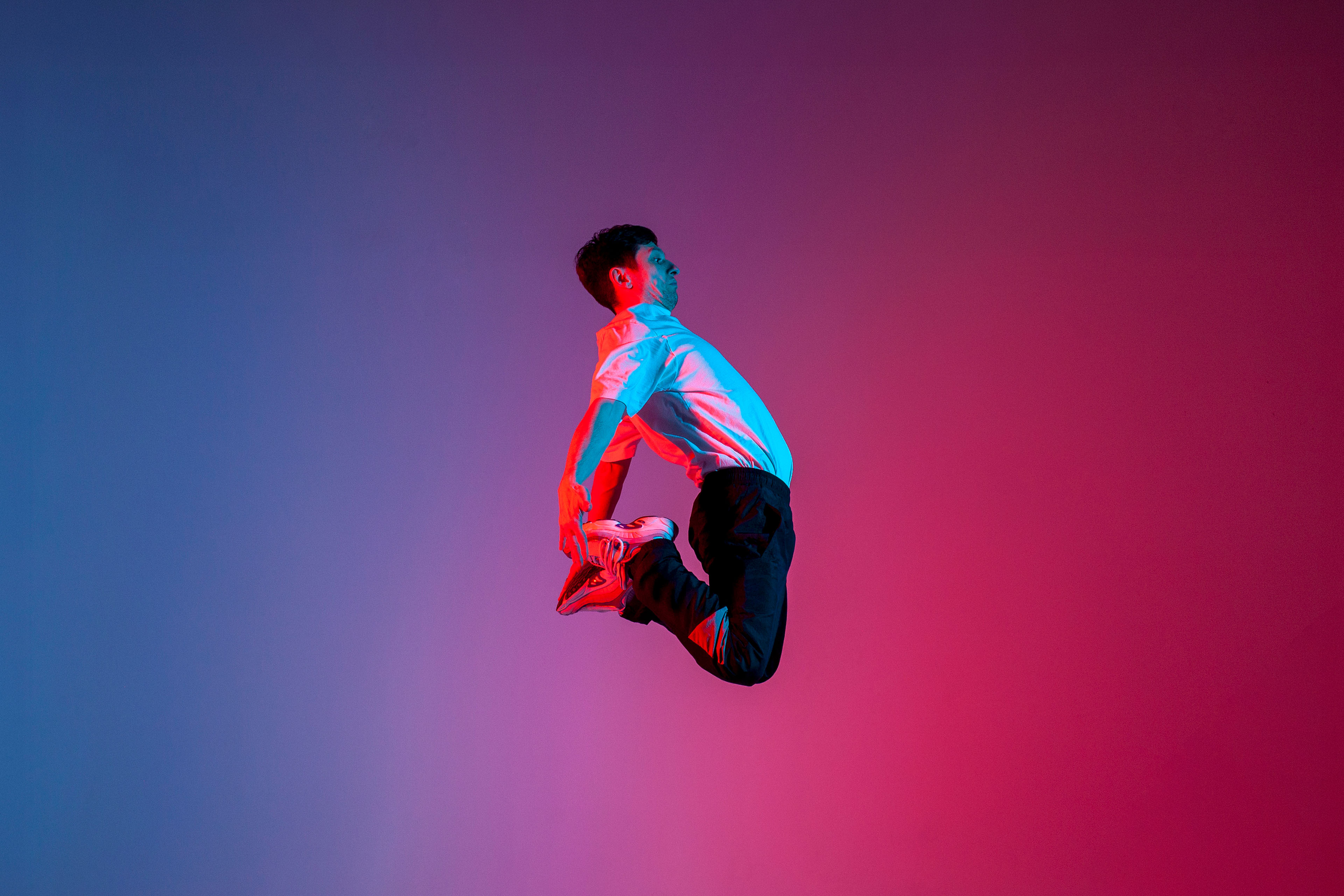 Danser in een sprong waarbij hij achter zich met zijn handen zijn voeten vastpakt tegen een blauw-roze achtergrond