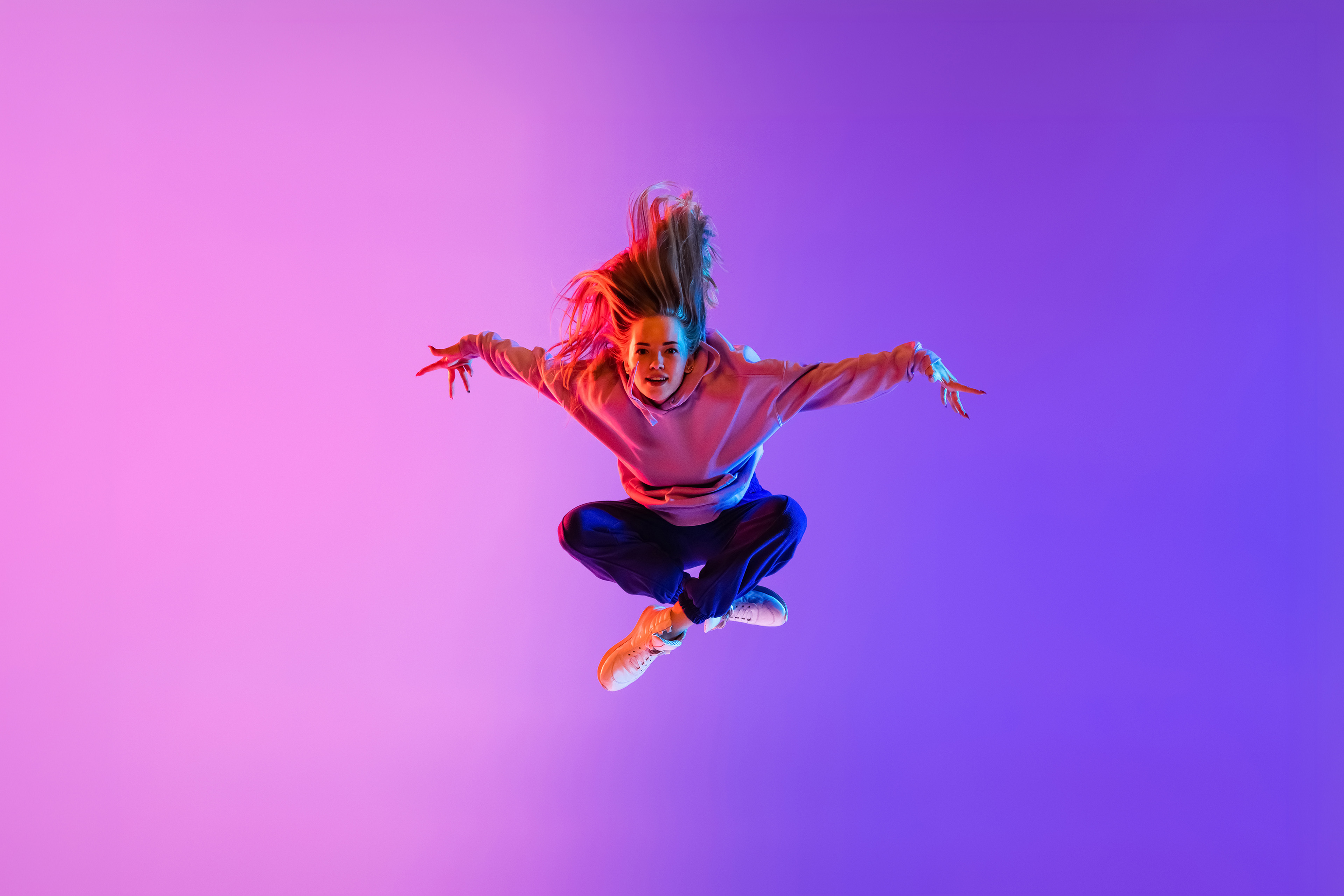 Danser springend in de lucht met uitgestrekte armen en benen in kleermakerszit, tegen een roze-paarse achtergrond