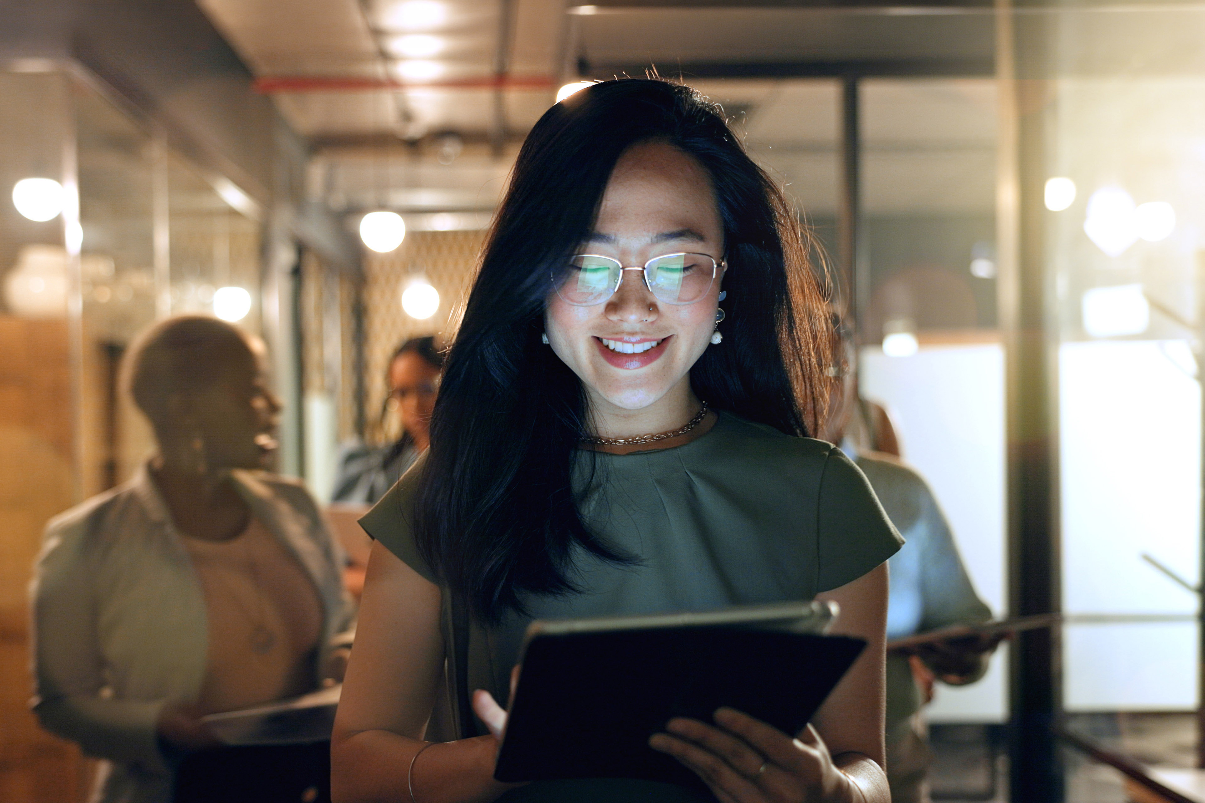 Persoon die door een kantoorgang loopt en iets bekijkt op een tablet, met collega’s op de achtergrond