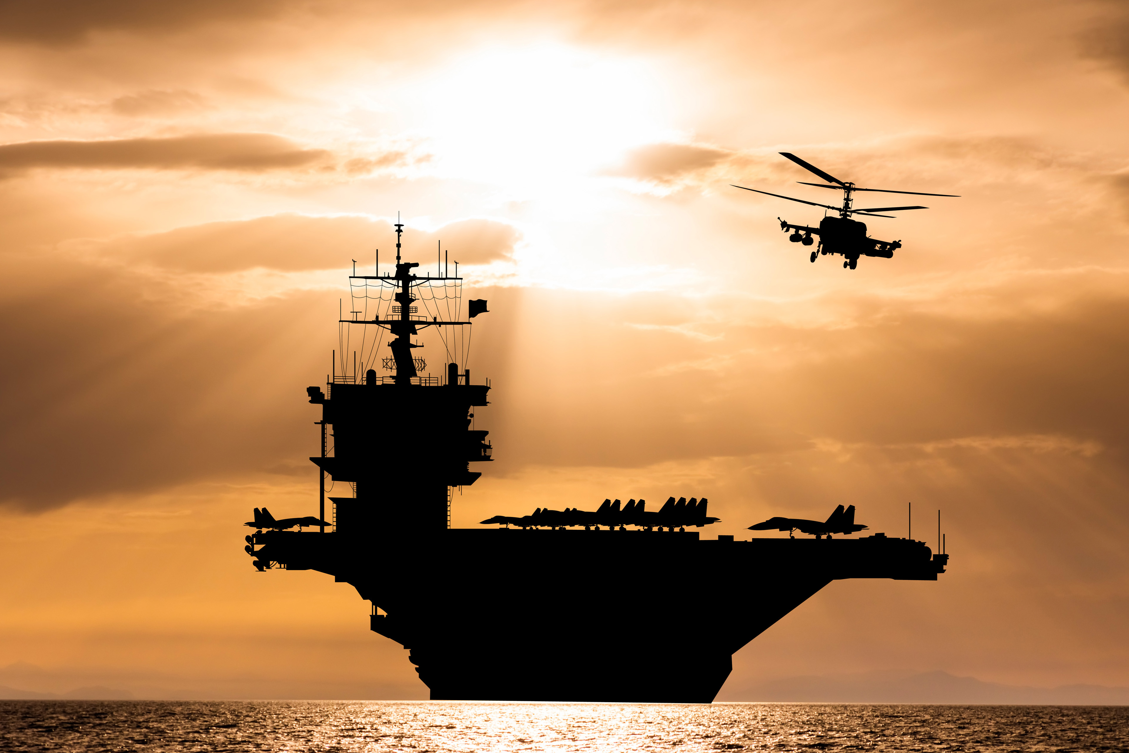 Silhouet van een vliegdekschip op zee met een helikopter bij zonsondergang.