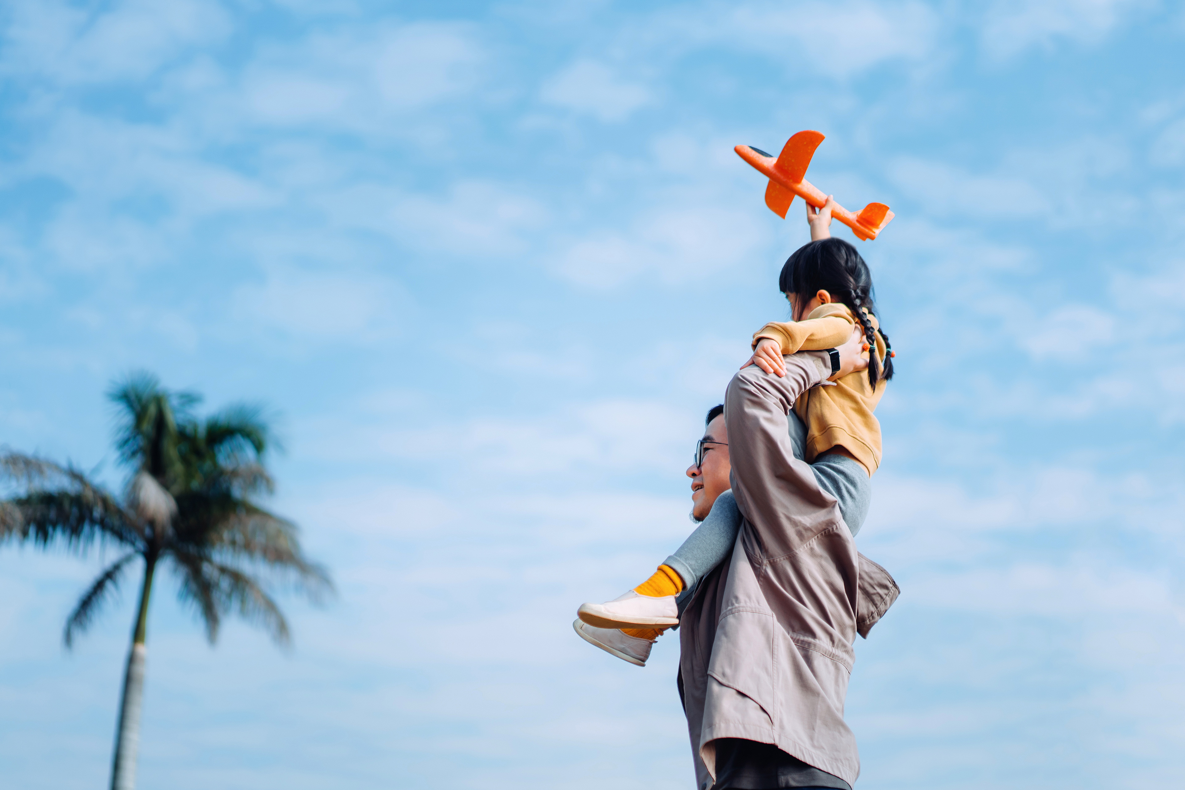 Volwassene draagt een kind dat een oranje speelgoedvliegtuig vasthoudt tegen een blauwe lucht.