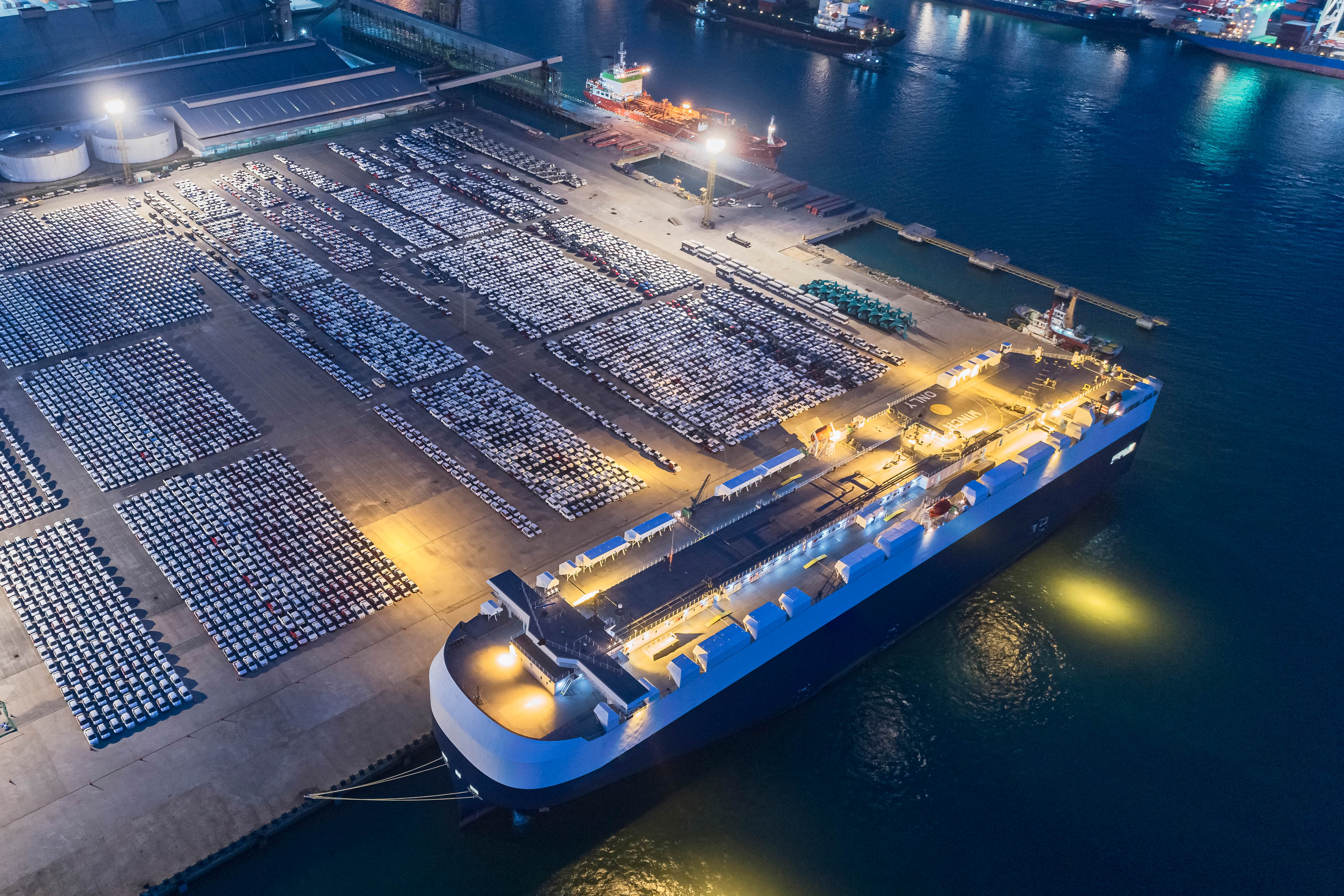 Luchtbeeld van auto’s opgesteld in een haven naast een groot vrachtschip ’s nachts
