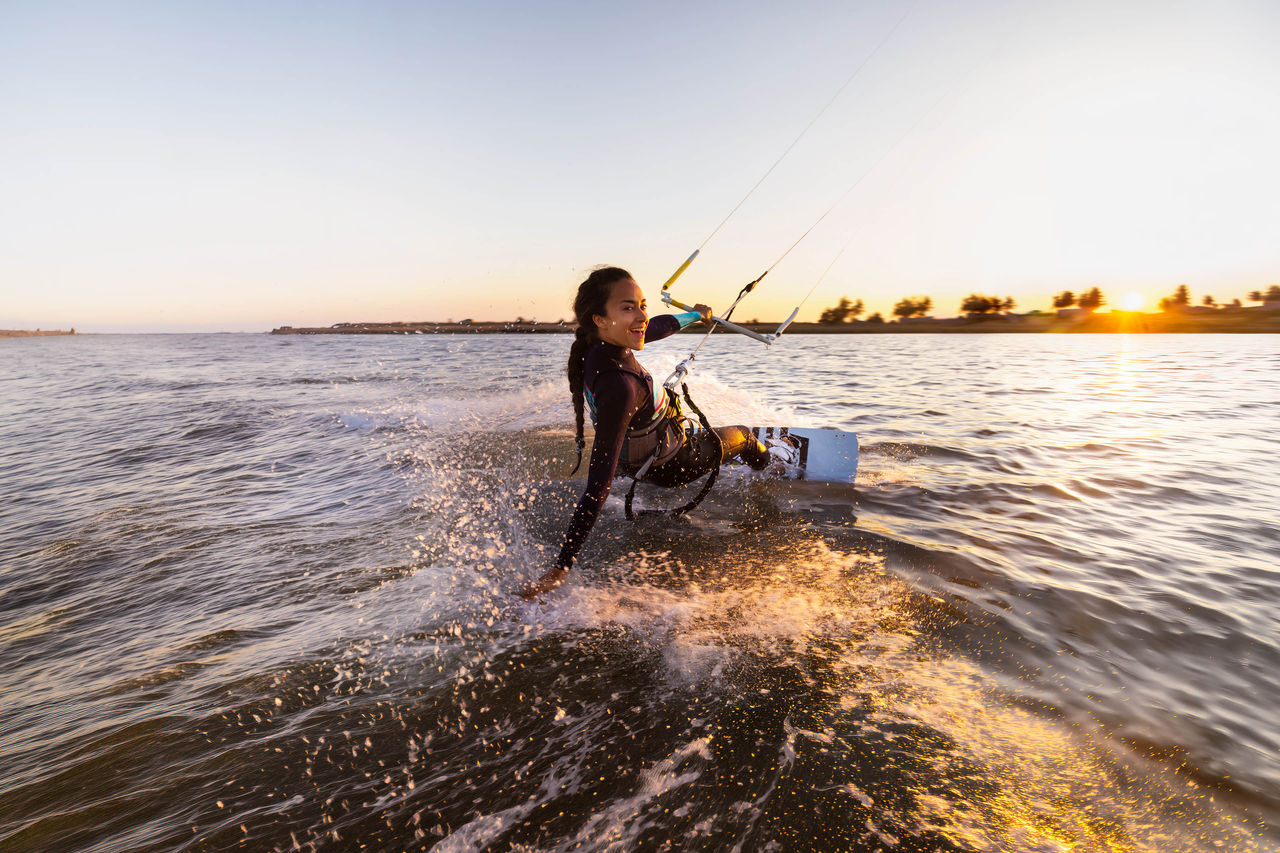 Kite surfing girl in swimsuit on board in blue sea riding waves with water splash on sunset. Recreational activity, water sports, action, hobby and fun in summer time. Kiteboarding sport.