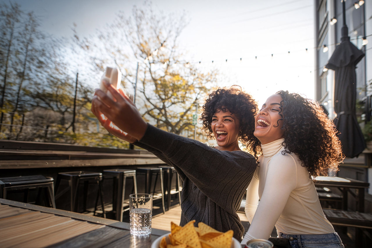 Young woman clicking selfie using an instant digital camera at a coffee shop. Two friends sitting with coffee cups on table having fun with a camera.