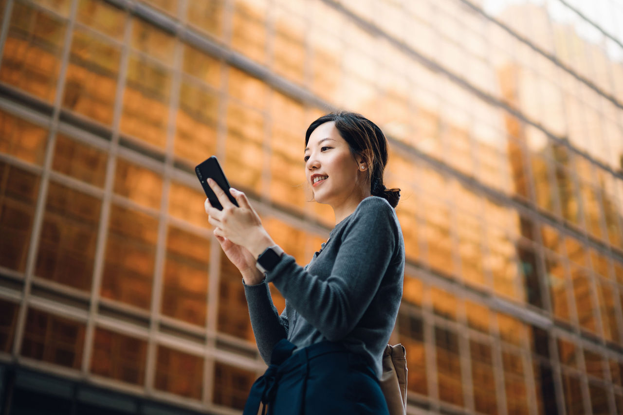 Low angle portrait of young Asian businesswoman managing online banking with mobile app on smartphone on the go. Transferring money, paying bills, checking balance in the city against corporate skyscrapers. Making business connections throughout the city