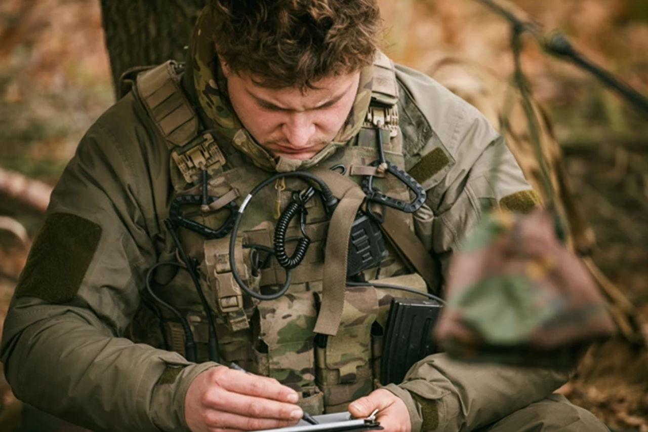 Soldier in camouflage uses a tablet while sitting outdoors in a wooded area.