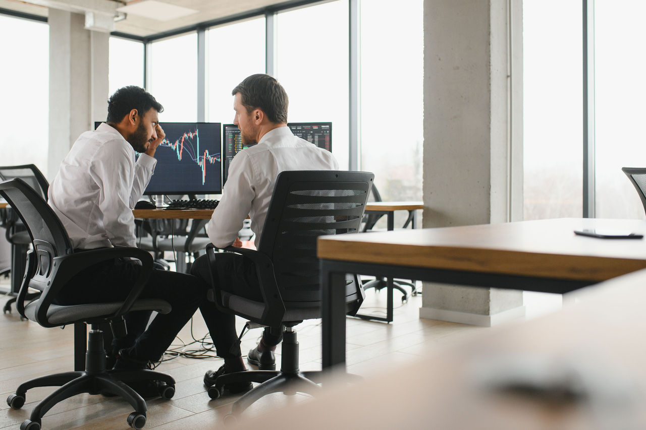 Two men traders sitting at desk at office together monitoring stocks data candle charts on screen analyzing price flow smiling cheerful having profit teamwork concept.