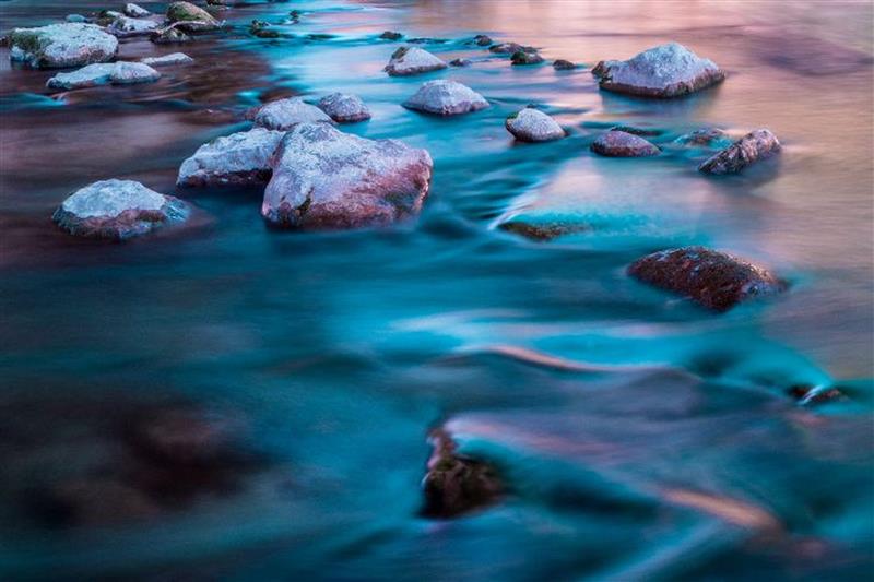 Fließender Bach mit Steinen bei Langzeitbelichtung, weiches Wasser in Blau‑ und Türkistönen.