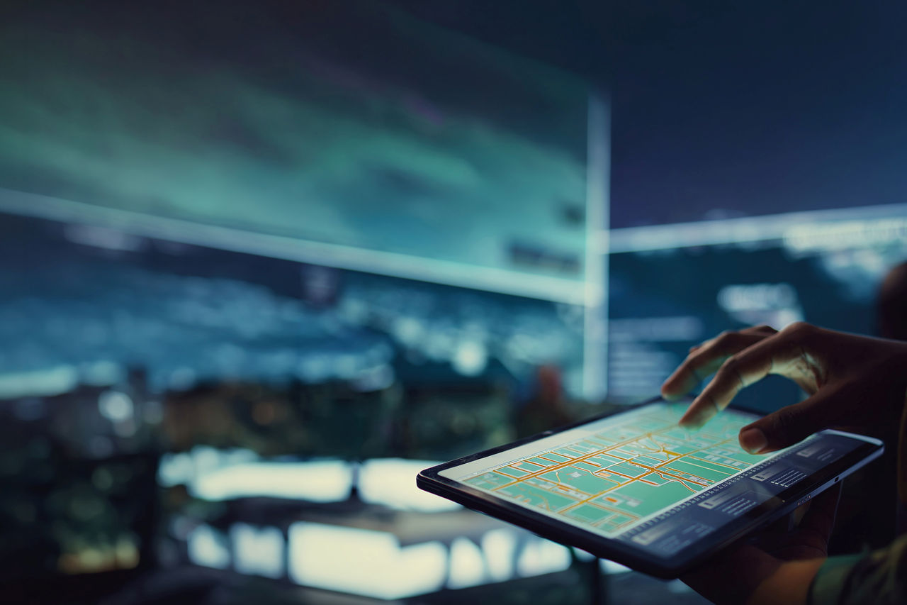 Person bedient ein Tablet mit digitaler Stadtkarte in einem Kontrollraum mit mehreren Datenbildschirmen.