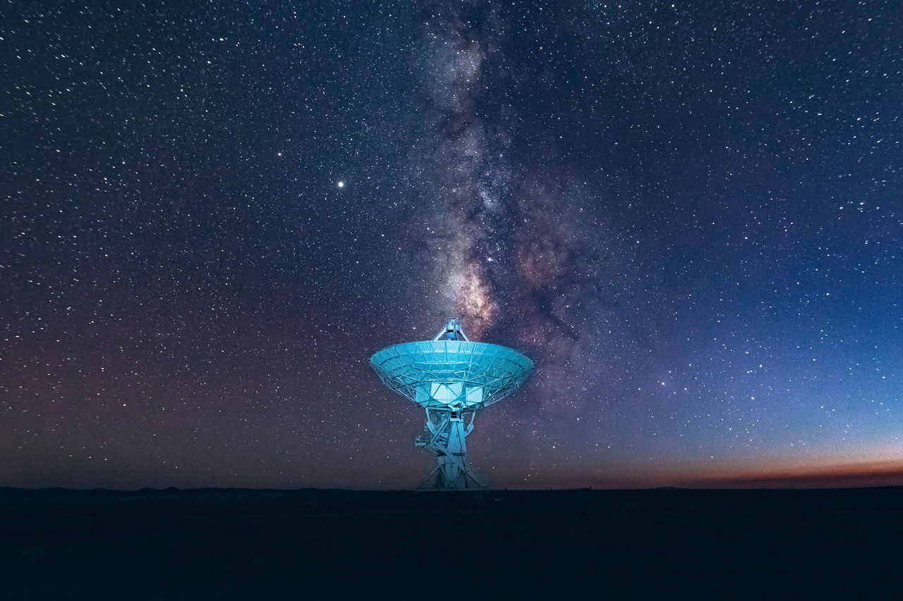 Große Satellitenschüssel unter dem Sternenhimmel, die Milchstraße leuchtet über einer stillen Landschaft.