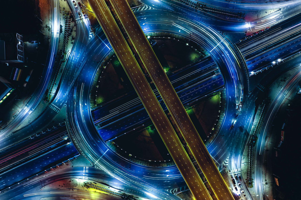 Moderne Straßenkreuzung bei Nacht aus der Luft, mit hellen Lichtspuren und dynamischem Verkehrsfluss.