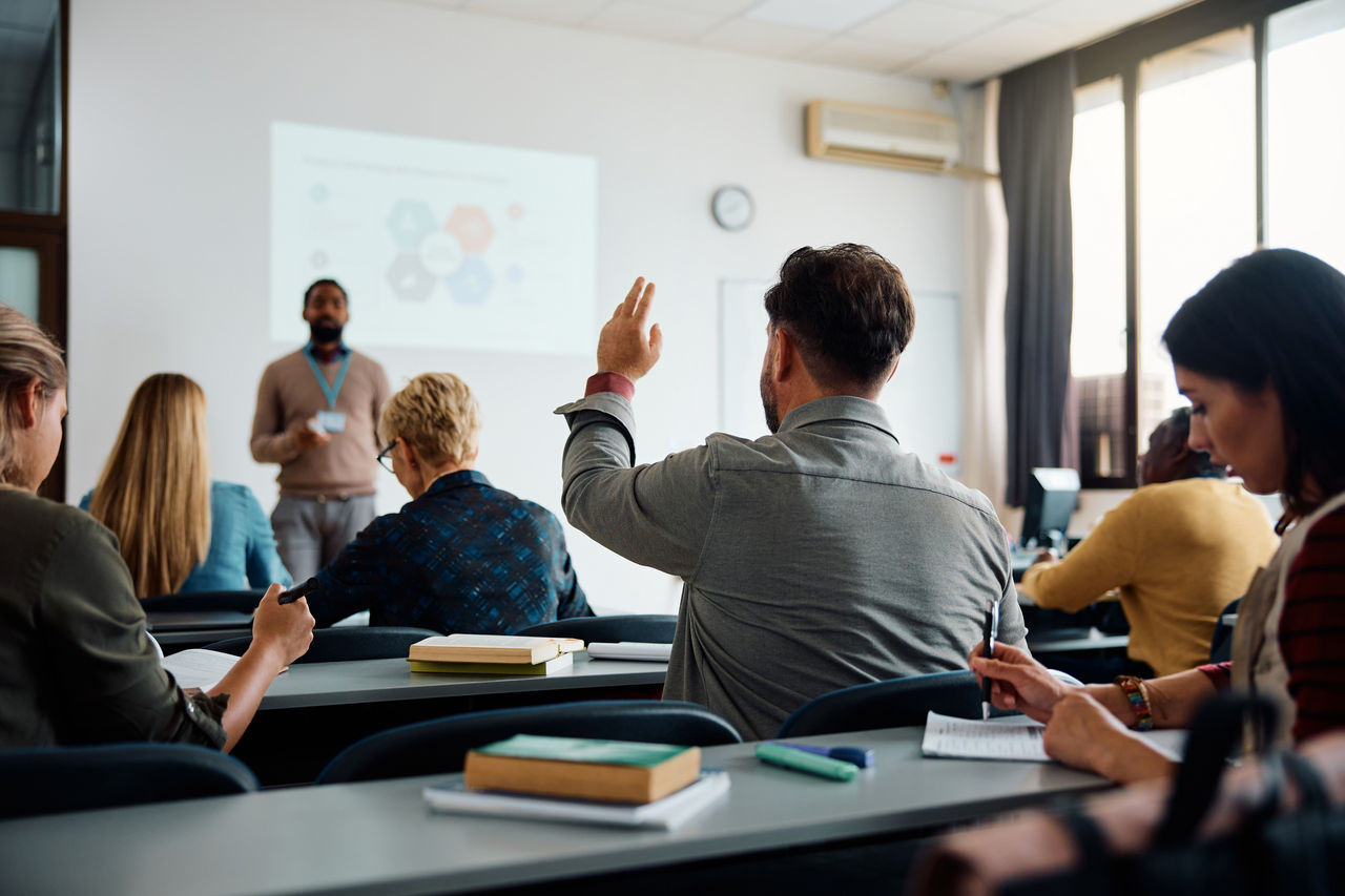 Seminarsituation mit Vortrag und Teilnehmenden, eine Person hebt die Hand für eine Frage.