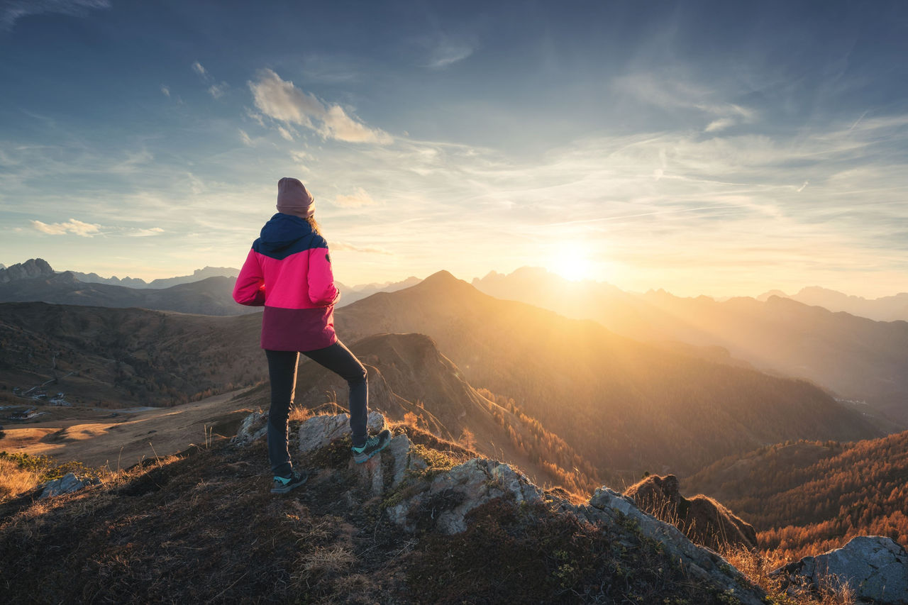 Person steht auf einem Berggipfel bei Sonnenaufgang und blickt über Täler und entfernte Hügel im goldenen Licht.