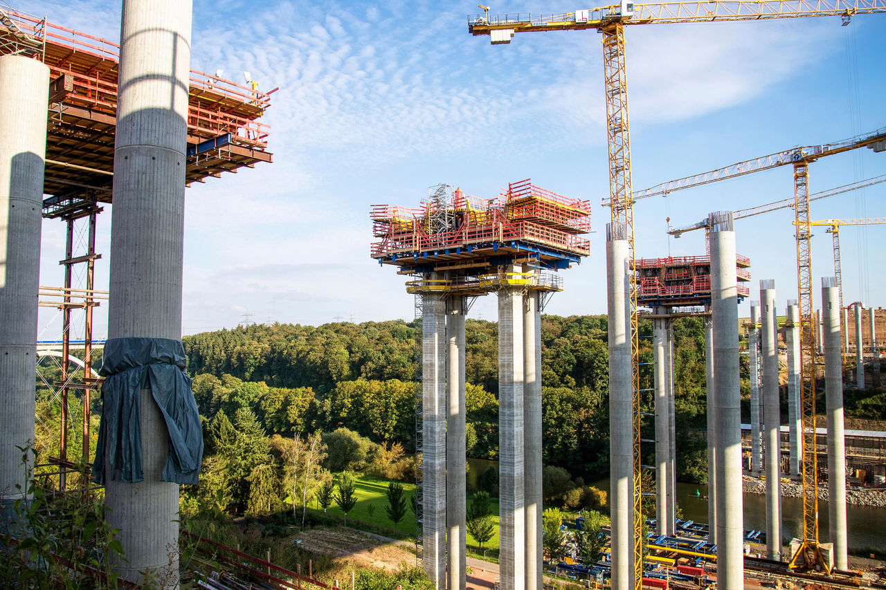 Großbaustelle einer Brücke mit hohen Betonpfeilern, Kränen und Gerüsten in grüner Landschaft.