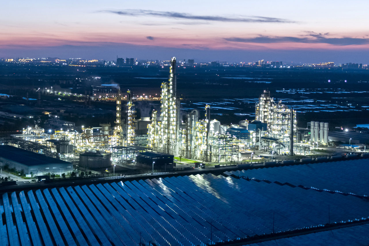 Beleuchtete Industrieanlage bei Dämmerung mit Solarmodulen und Stadtlichtern im Hintergrund.