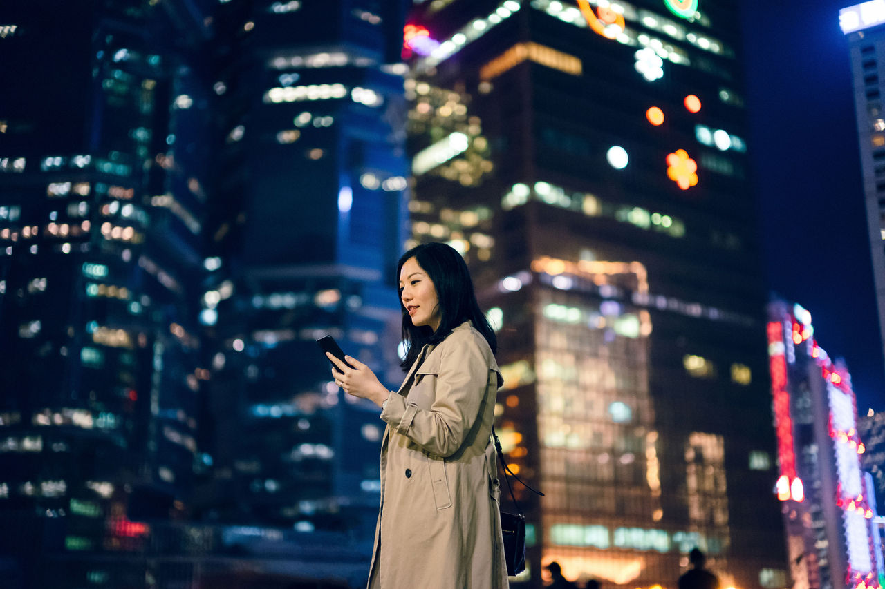 Confident young businesswoman using smartphone against Hong Kong cityscape with contemporary skyscrapers in financial district