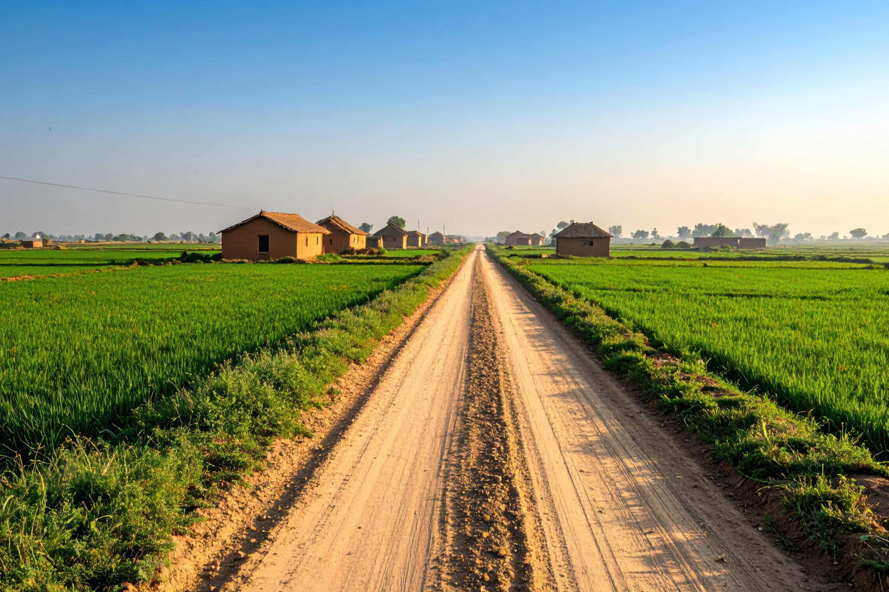 Dusty road stretches through a serene rural landscape of green fields and small village houses under a clear blue sky